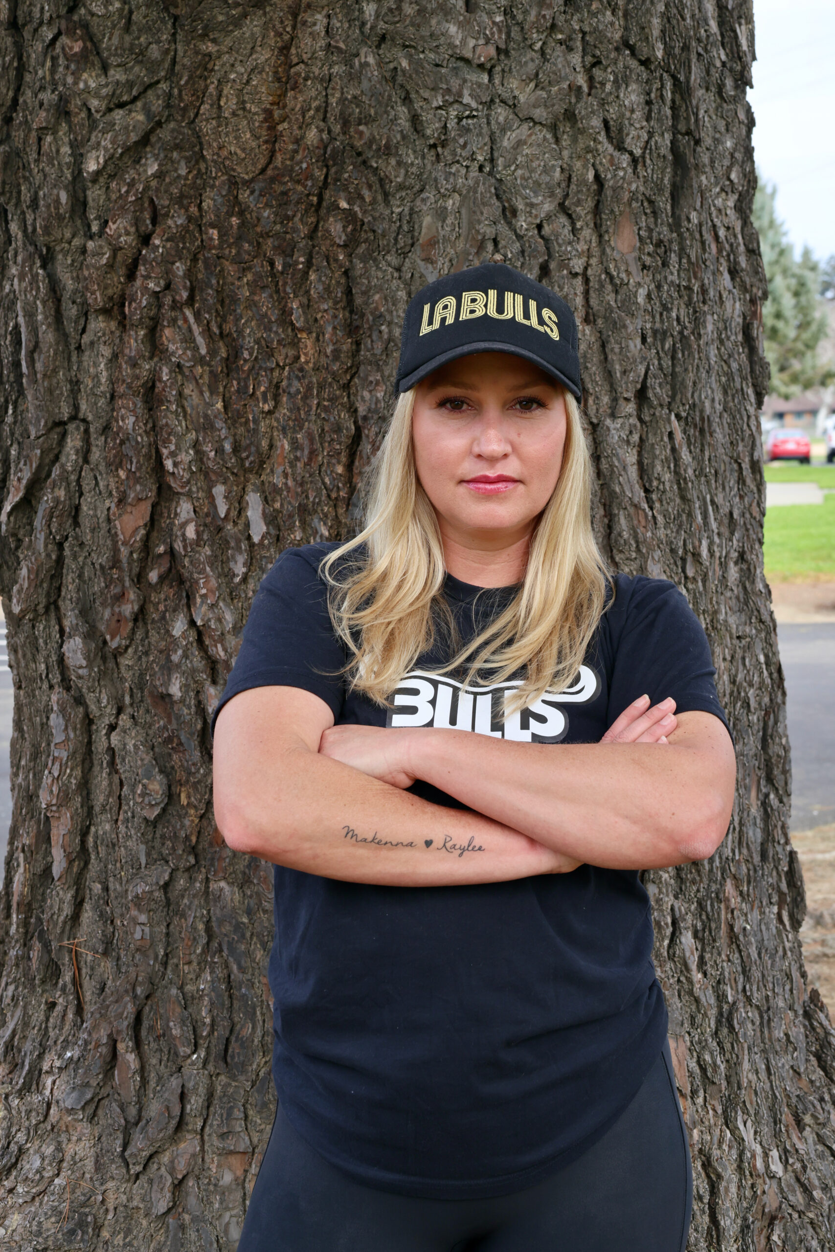 Young Woman with blond hair in a black hat, standing in front of a tree with her arms crossed