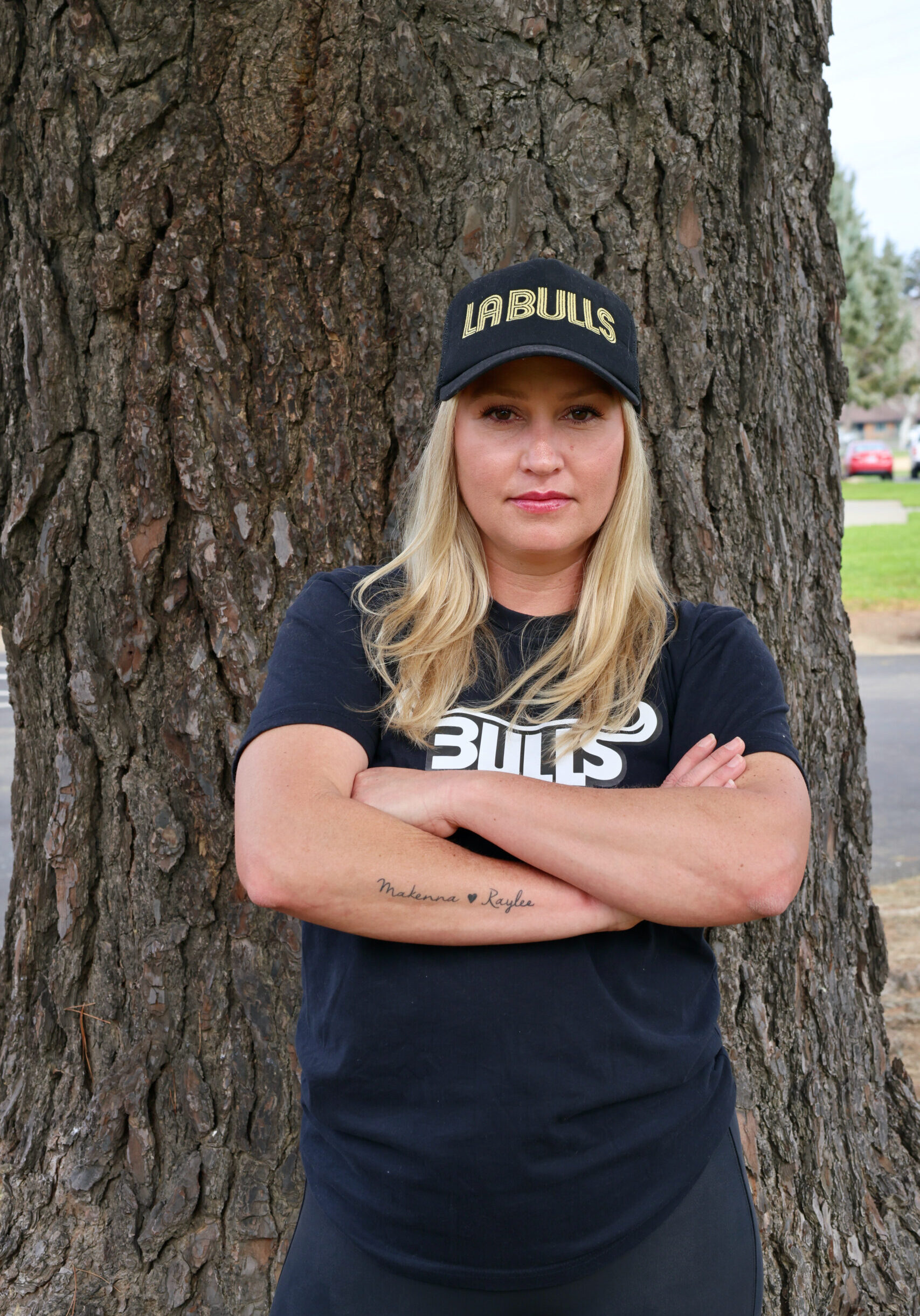 Young Woman with blond hair in a black hat, standing in front of a tree with her arms crossed